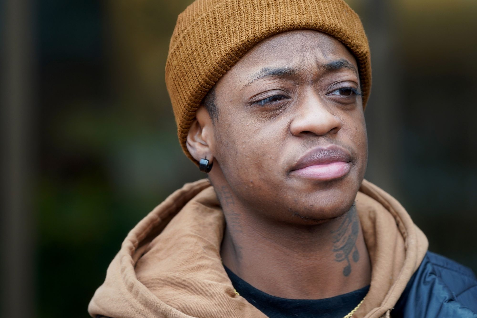 A Black man with a dark yellow beanie, an earring and a neck tattoo looks to the side. 