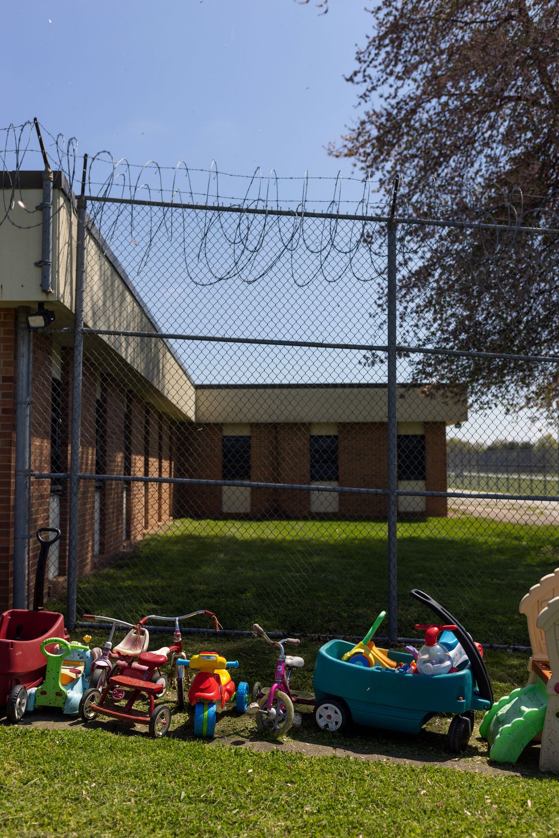 A photo shows toys and children’s bikes on the grass in front of a barbed wire fence and a brick prison building.