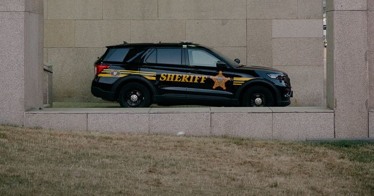 An empty black sheriff's vehicle, with the word "Sheriff" printed on the side in yellow paint, is parked at a beige-colored building with two columns on either side.  