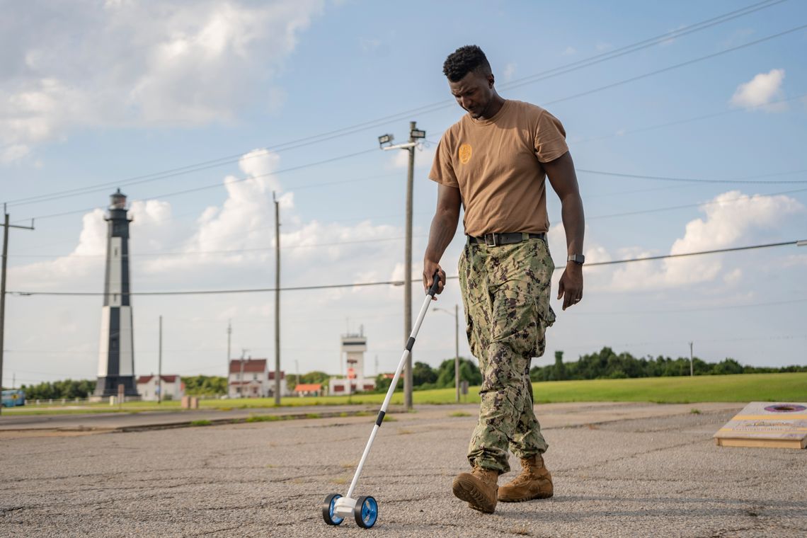 Master of Arms Vincent Montague, a Black man, arranges a cornhole board set in an outdoor parking lot. 