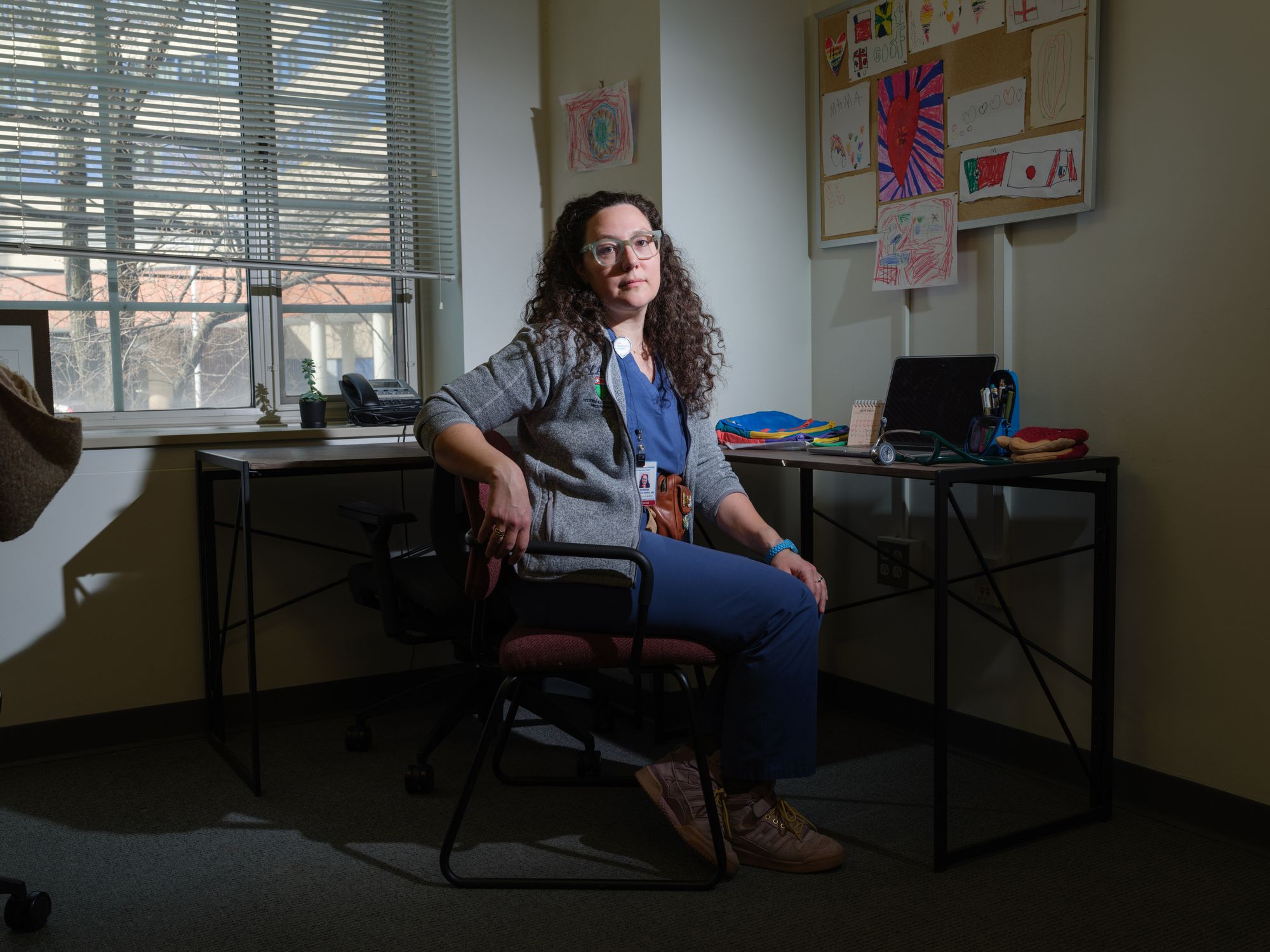 A White female doctor, wearing glasses, a gray jacket and blue scrubs, sits at her desk. Papers of children's hand drawings hang on the wall.