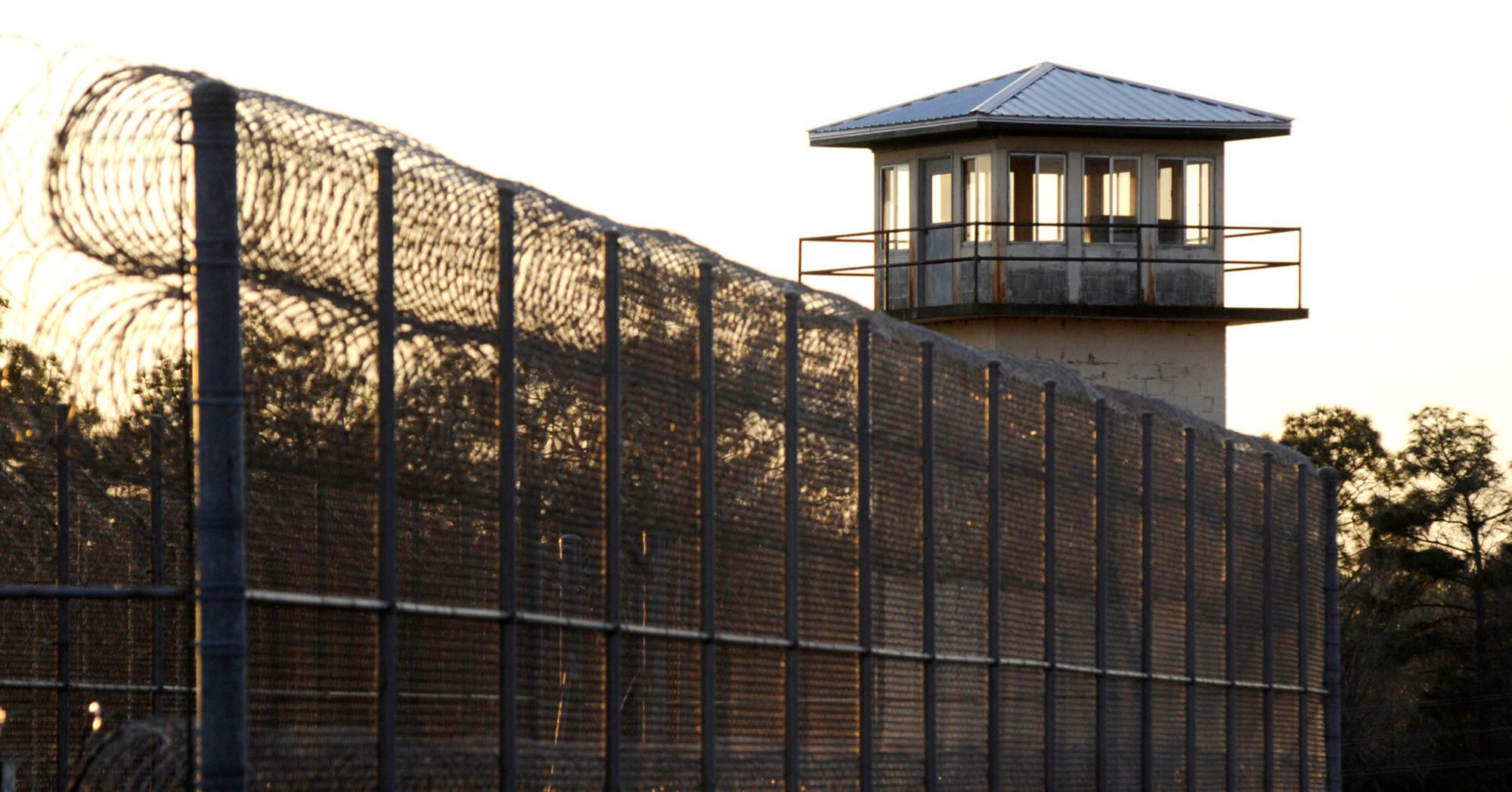 A photograph shows barbed wire and a prison tower at Holman Prison at sunset. 