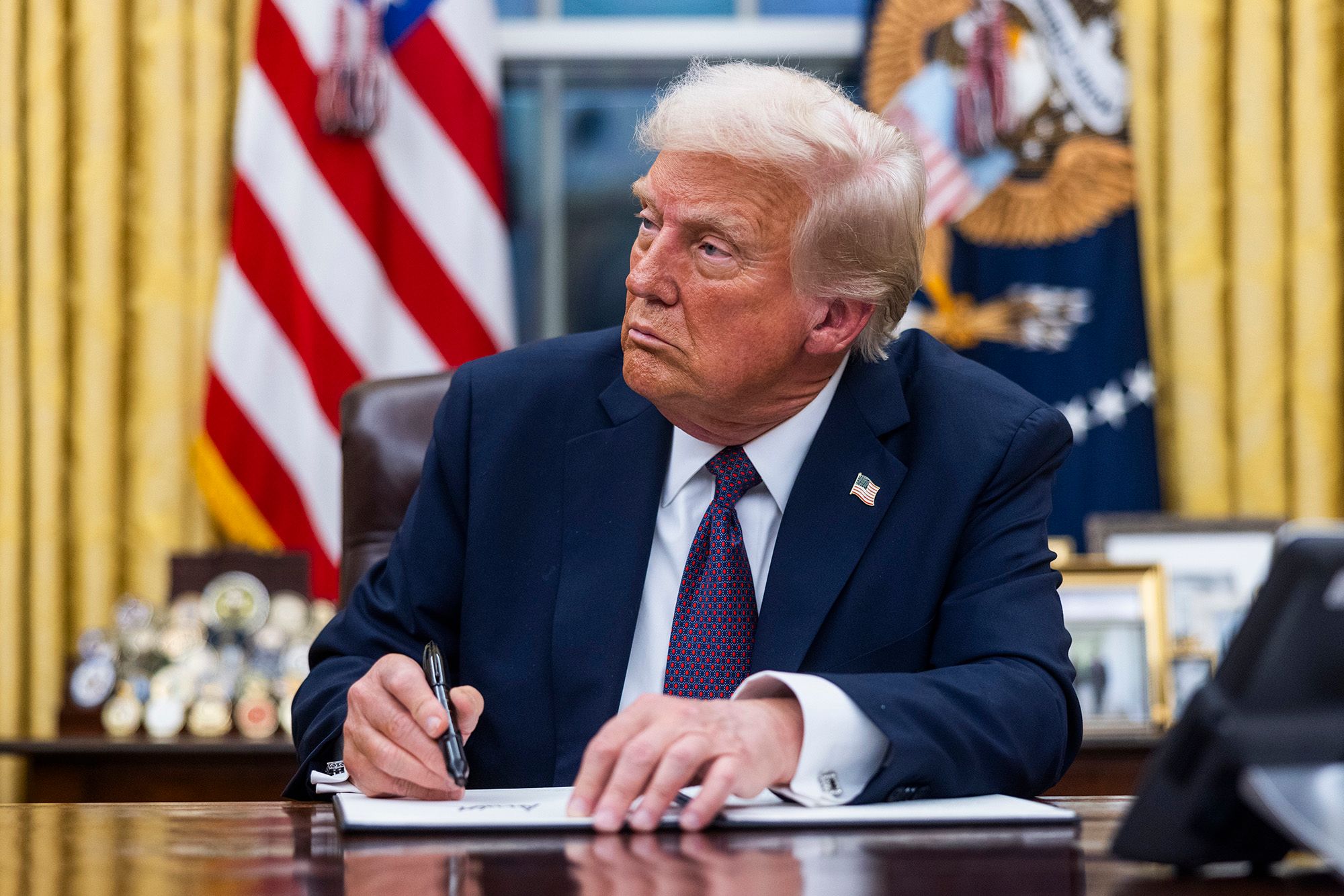 Trump, a White man in a navy suit and a red and navy tie, signs a document while sitting in the Oval Office. 