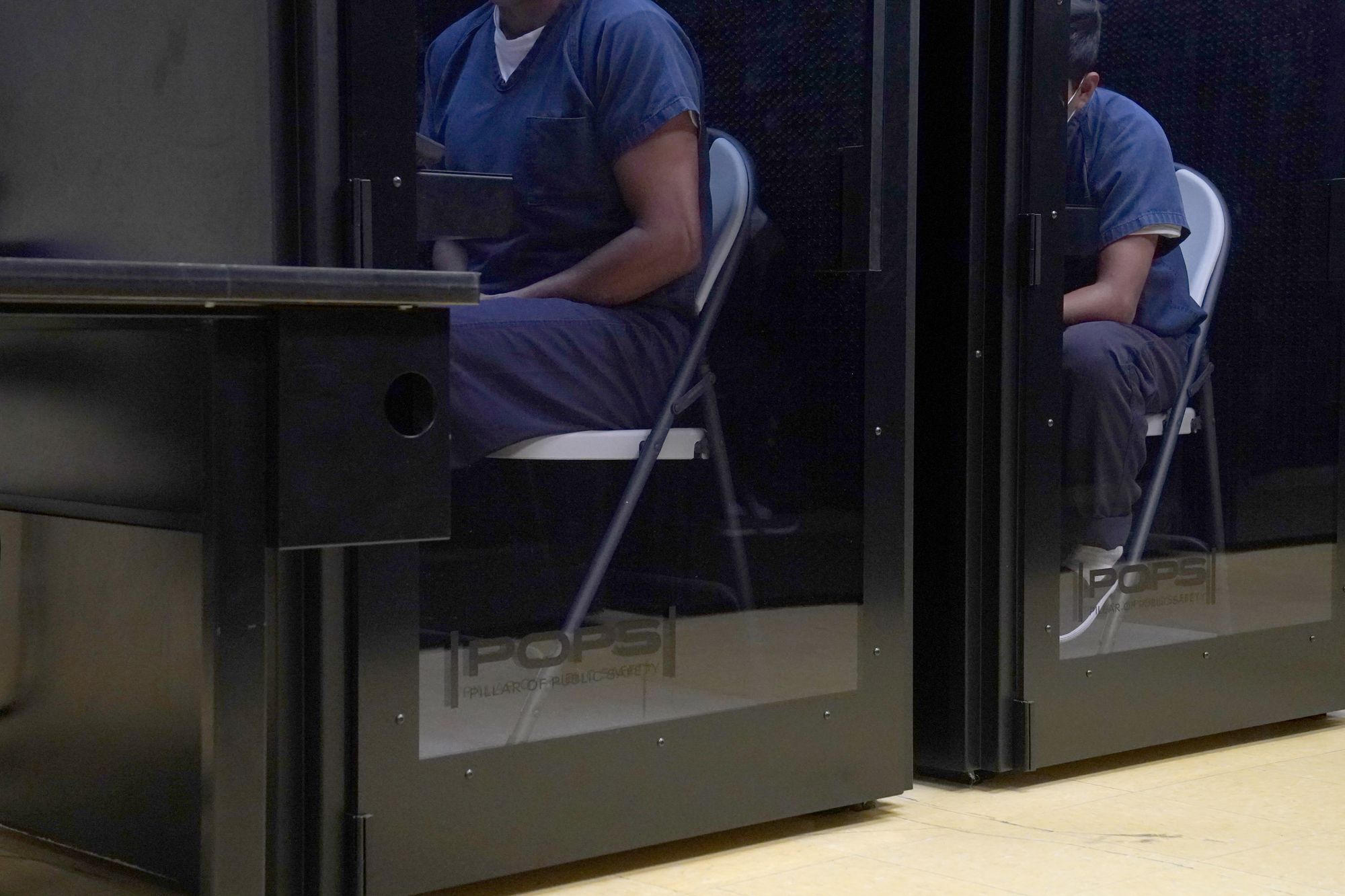 Two detainees in blue uniforms, whose faces are not showing in the photograph, sit in privacy booths talking to their attorneys or asylum officers. 