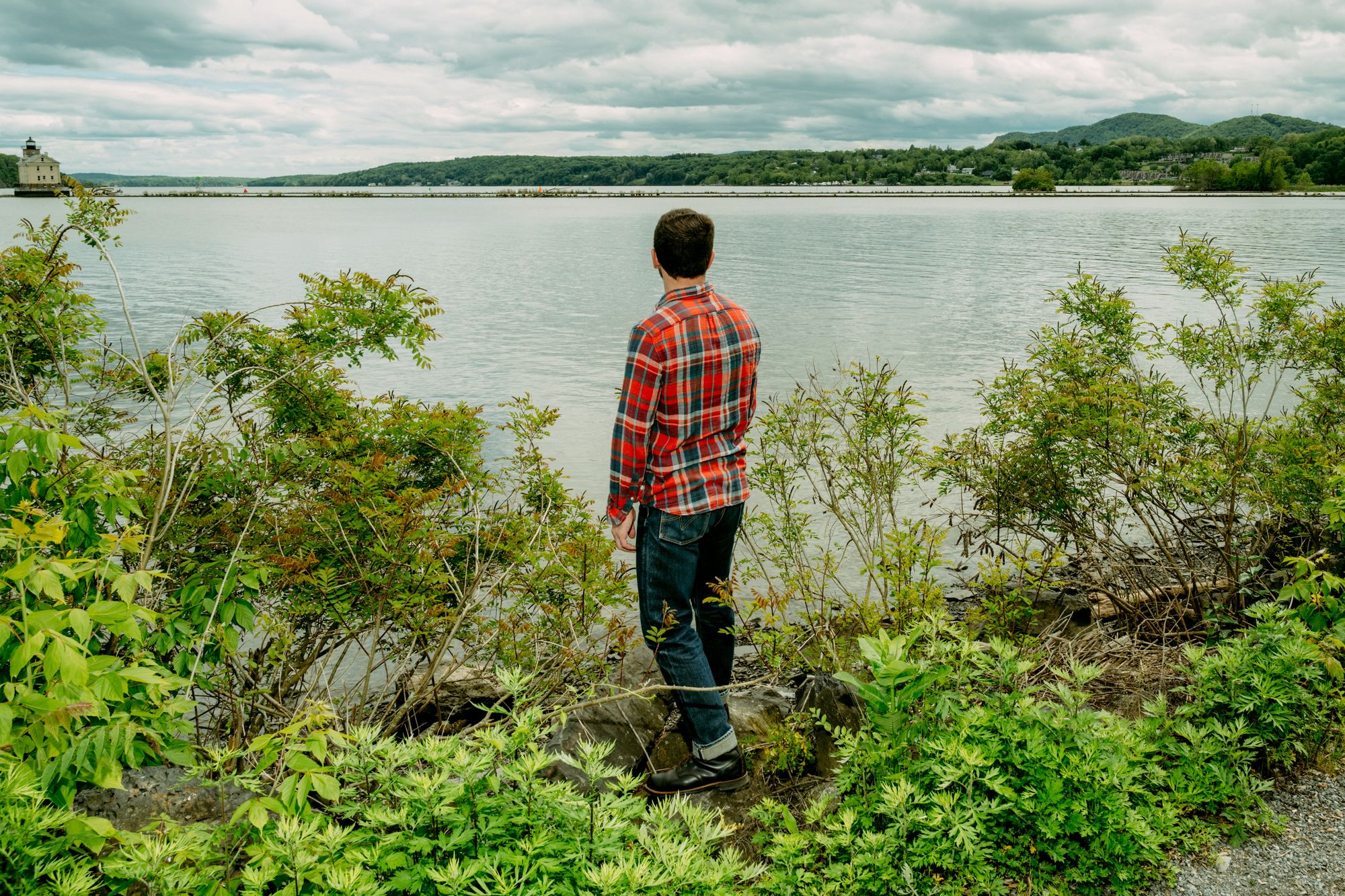 Michael McCallion, a man with light-toned skin and short dark brown hair, wears an orange plaid long-sleeved shirt and jeans while standing near a lake.