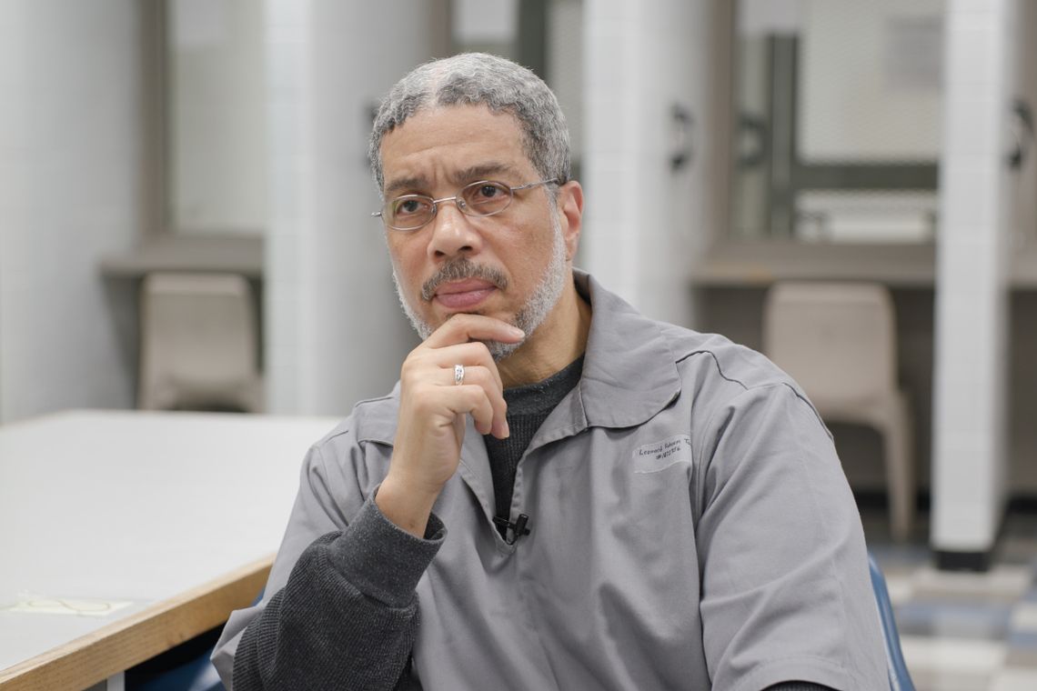 A Black man, wearing glasses and a gray prison uniform, sits in a visiting room. He places his hand under his chin as he looks outward. 