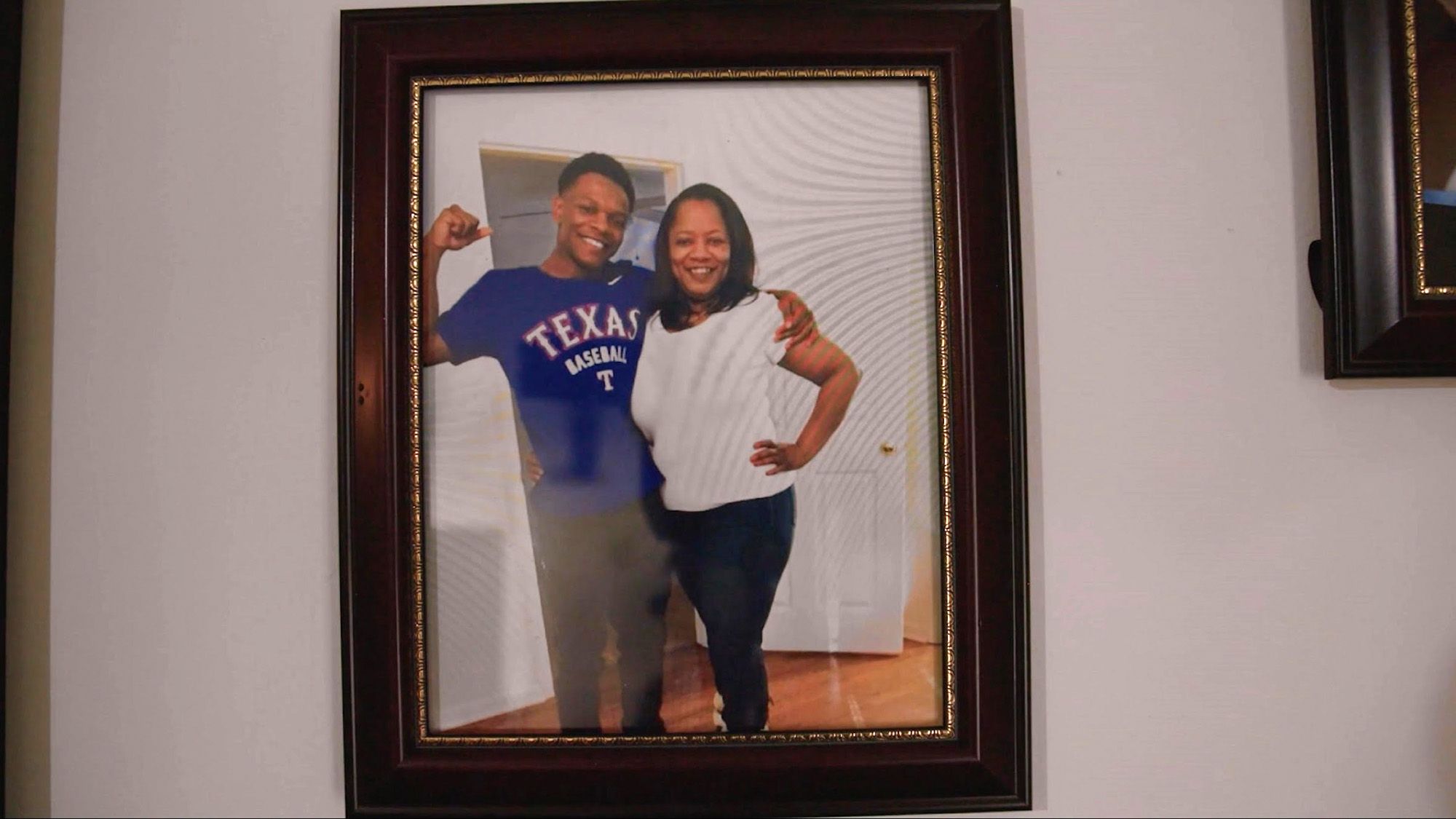 A framed picture of Angela Williams, a Black woman wearing a white top and black pants, smiling and standing with her son Ulysses, a Black man in a blue T-shirt that reads “Texas baseball” and black pants. 