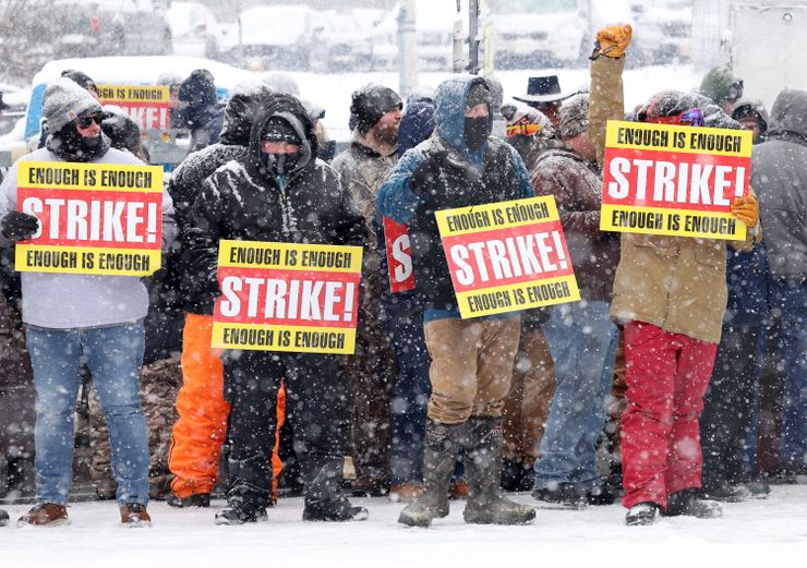 In the foreground, four people wearing winter coats hold up signs reading "Enough is Enough, Strike!" as it snows.  A group of people stand behind them.  