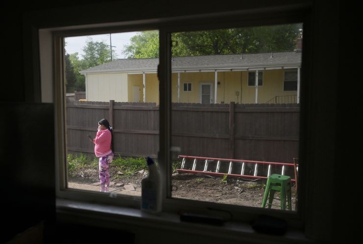A photo shows a woman with medium skin tone through a window frame, wearing a pink jacket and purple tie-dye leggings, looks at her phone while standing outside her home. 