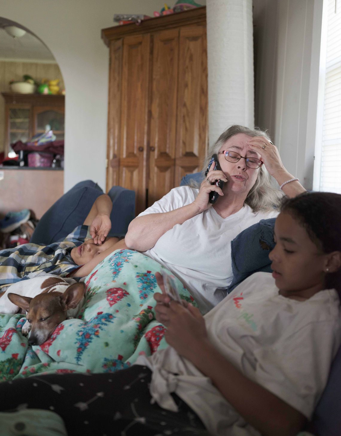 A White woman looks tired as she talks on the phone while sitting with her grandchildren. A young multiracial boy, lies to her right, and a young multiracial girl, sits to her left. 