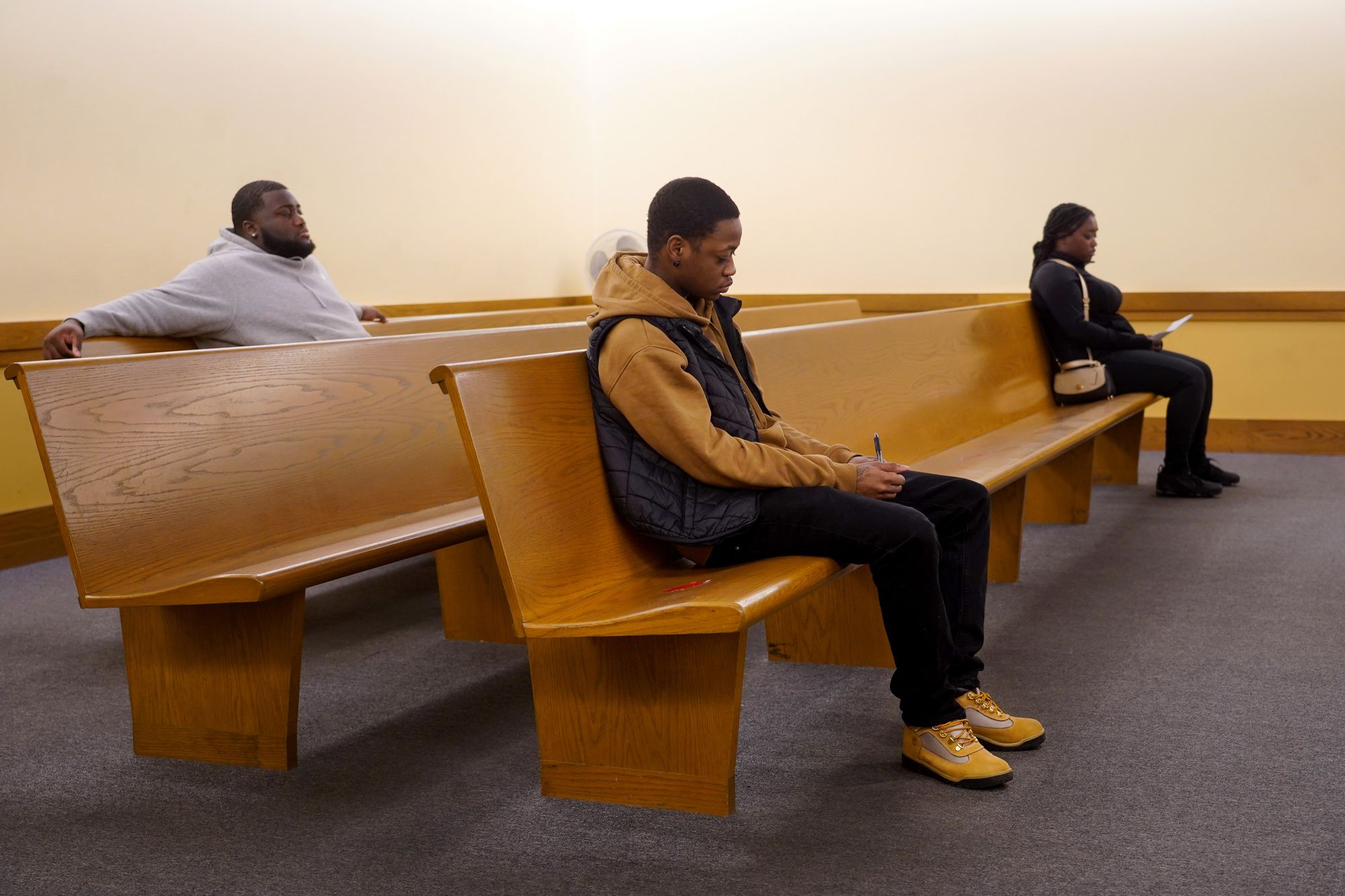 Three Black people sit widely spaced away from each other on courtroom benches. The man in the center of the picture, wearing a dark yellow hoodie and black vest, fills out a form. 