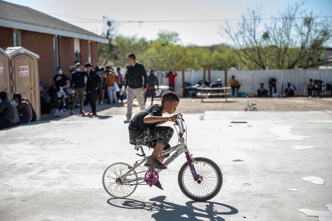 Andrew Cordero Bullain, a young boy with close-cropped dark brown hair and medium-toned skin, sits on a bicycle in the center of the photograph. Some people are gathered in the background at the respite center. 