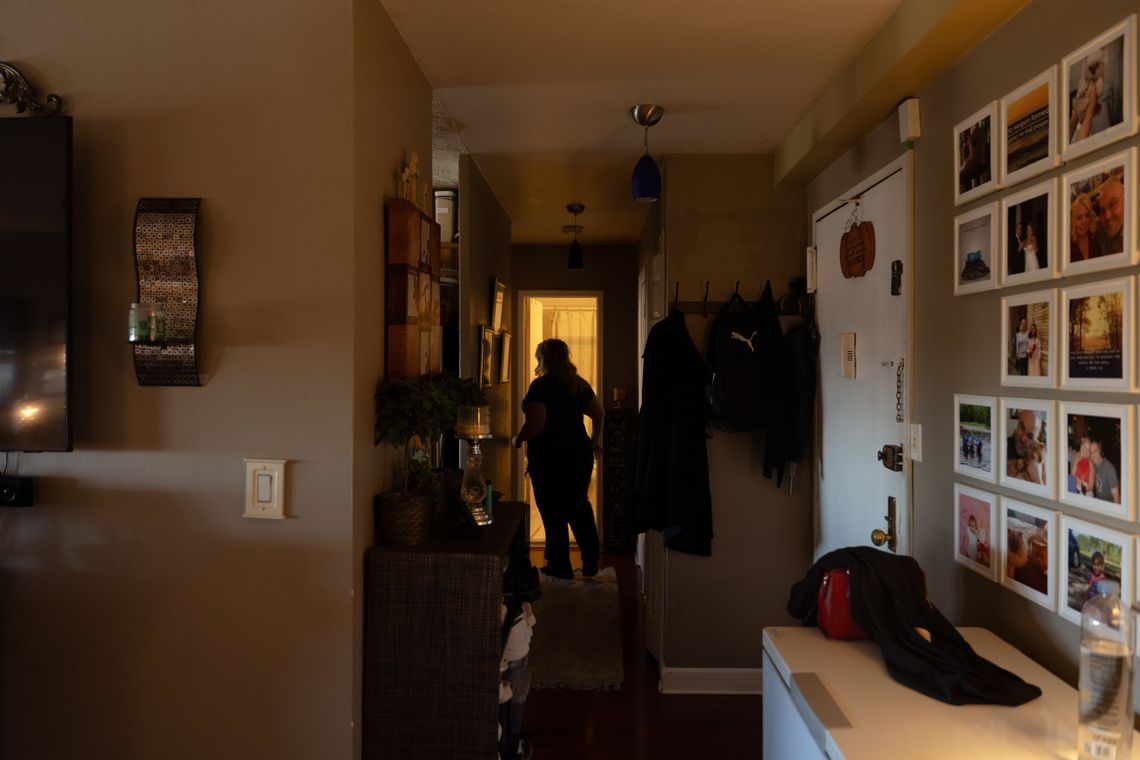 A photo shows the silhouette of Sandra standing in front of her bathroom door at the end of a short hallway. In the foreground on the left is a wall of family photos, a wall-mounted coat rack, and the front door. 