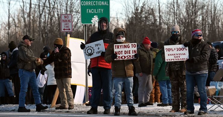 Protesters, wearing puffer jackets, beanies, gloves and pants, stand on the side of the road holding signs such as: "70% doesn't equal 100%," "Trying not to die in 2025," and "Essential not expendable." In the background, a road sign reads "Greene Correctional Facility." 