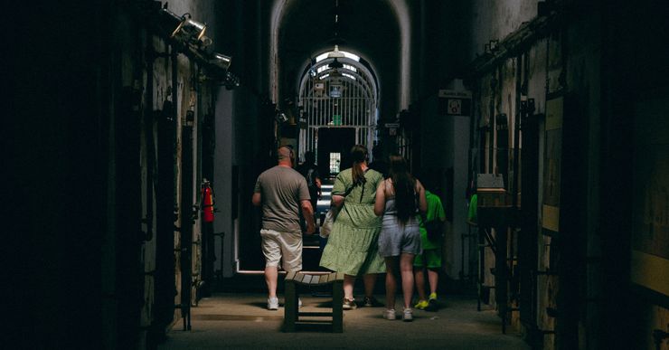 A group of people walk through a dark hallway with prison cells on either side.