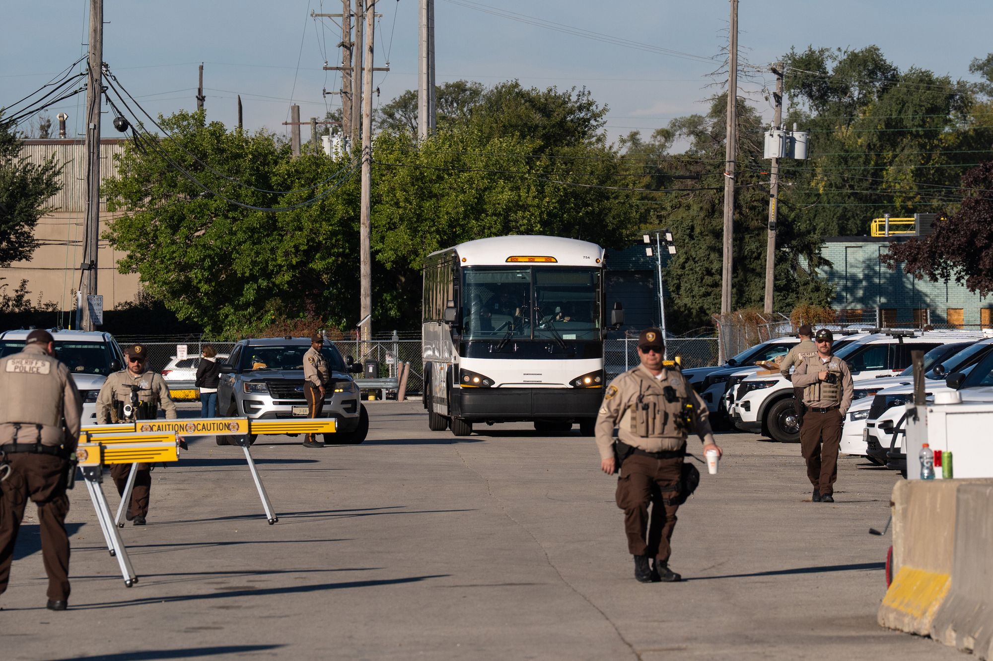 Sheriff’s officers in brown uniforms stand in front of a white bus in motion in a parking lot. 
