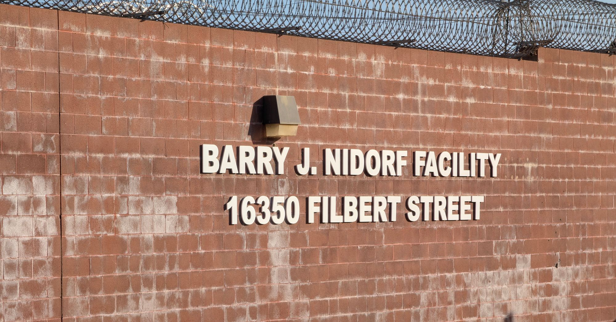 A photo shows a brick wall with barbed wire and a sign that reads, "Barry J. Nidorf Facility, 16350 Filbert Street." 