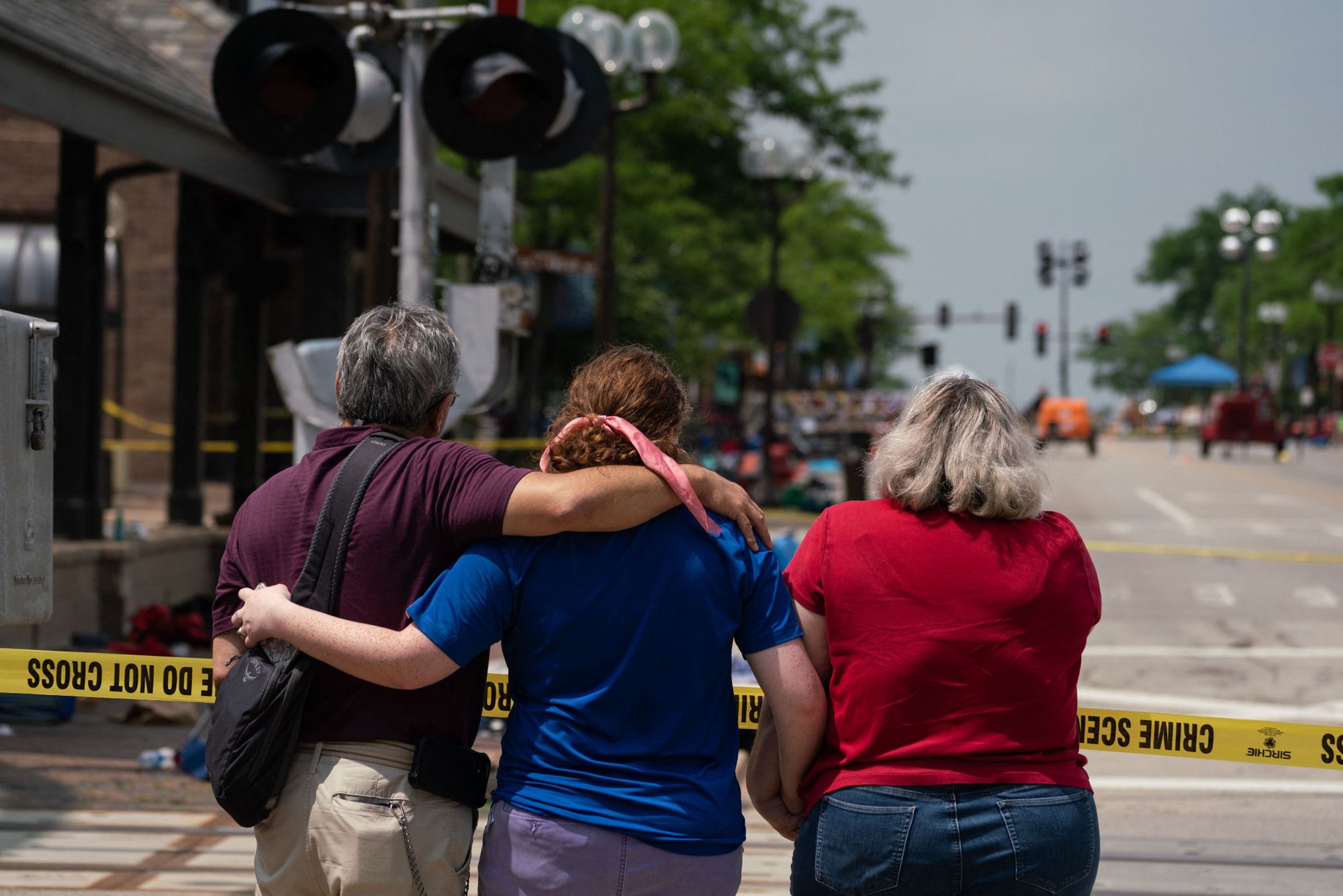 A man with gray hair, wearing a plum-colored t-shirt, has his arm around a woman with reddish-brown hair, wearing a blue t-shirt. She is holding hands with a woman with blonde hair, wearing a red t-shirt.  They all stand in front of police tape, looking down the street.  