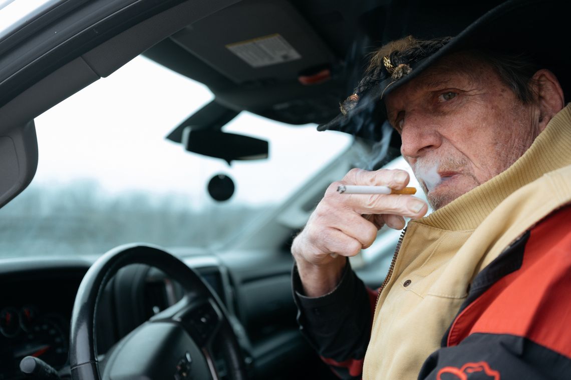 A photo shows George Hall, a White man in a beige vest and a red-and-black jacket, smoking a cigarette while sitting in the driver's seat of a car.