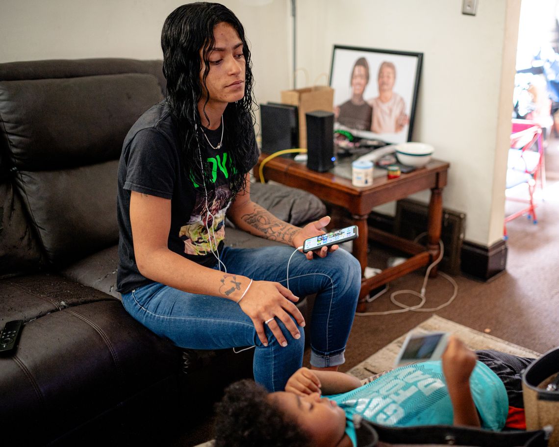 A photo of a young Black woman sitting on a couch while listening to audio from her phone through wired headphones. A Black boy with a turquoise shirt is laying on the table in front of her. 