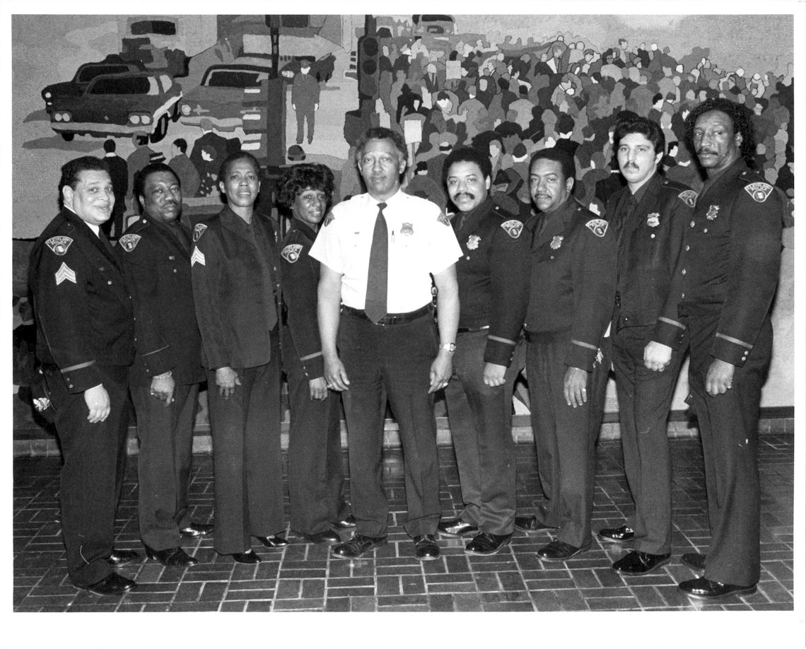 A black-and-white photo of 10 uniformed police officers standing in a line. Nine wear matching dark uniforms, and one in the center wears a lighter uniform shirt. A mural in the background depicts a bustling urban scene.