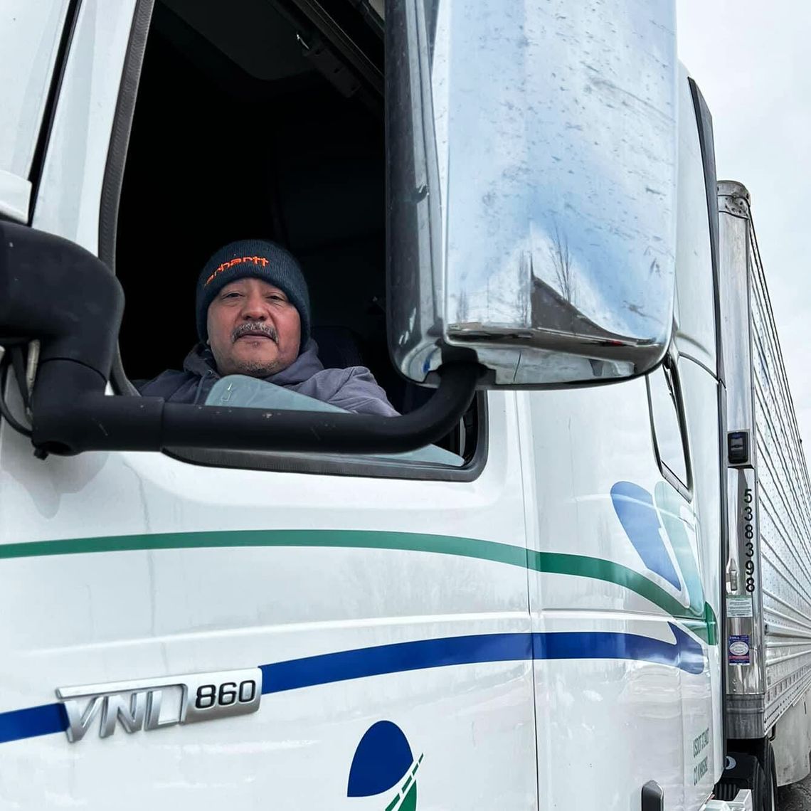 A man with a medium skin tone and a gray mustache sits in the driver’s seat of a semitruck. He is wearing a black beanie with orange text. 