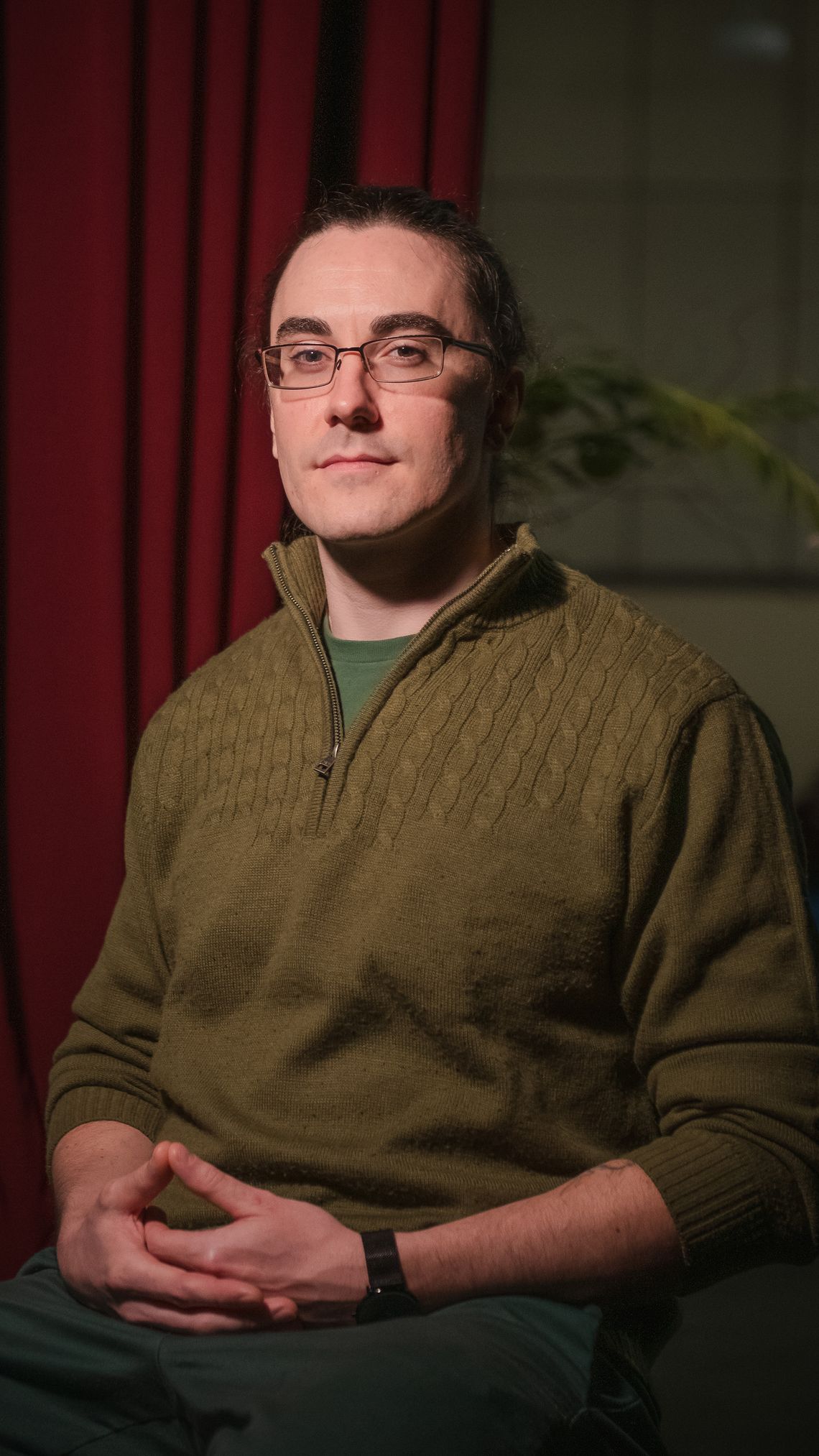 A White man wearing an army-green quarter-zip sweater sits with his hands together on his lap. Behind him is a dark red curtain. 