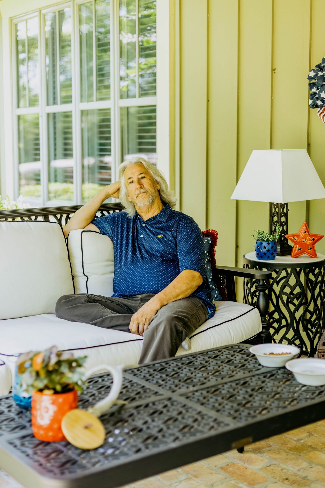 A photo of John Cook, a White man with gray shoulder-length hair, a gray beard and a blue polo shirt. Cook is sitting on a couch on a patio while looking into the camera and resting his head on his hand. 