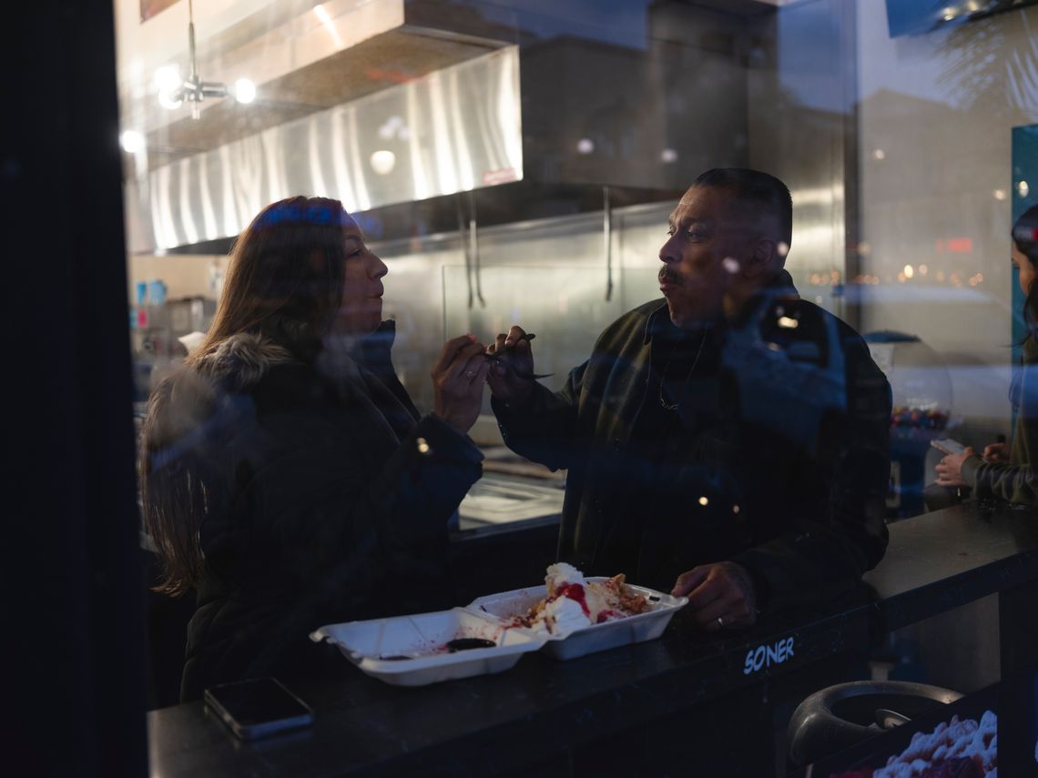 Silvia and Miguel are seen through the window of a funnel cake and churro shop, sharing a dessert.