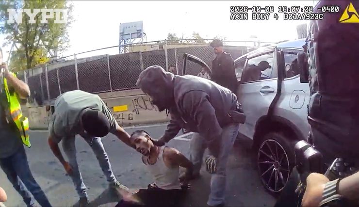 A video screenshot shows a dark-skinned man, wearing a white tank top and black pants, tilting his head as he sits on the floor. Two individuals standing next to him bend down to check on his condition. One person on the side is on the phone. Another person is exiting their car. A police officer stands in the foreground. 