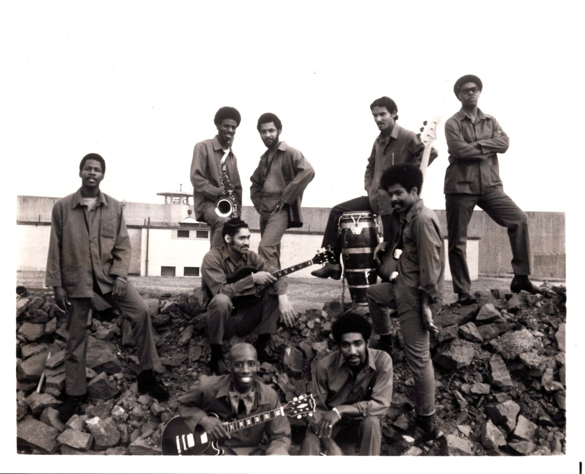 A photo of nine men wearing prison uniforms standing atop a pile of rocks at a prison. Some are standing at the ridge of the rockpile, one of them holding a saxophone and another, his left leg splayed over a bongo drum. In front of them sits a man with long sideburns and a guitar resting on his lap. On each side of him stand two men. In the foreground, one man with an afro sits smiling, his right elbow on his right knee. Just to the right sits a bald man, smiling, and holding a guitar. 