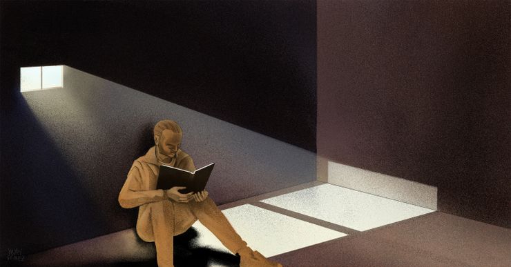 A prisoner sits inside a dark cell, reading a book. Light from a small window shines through the room. 