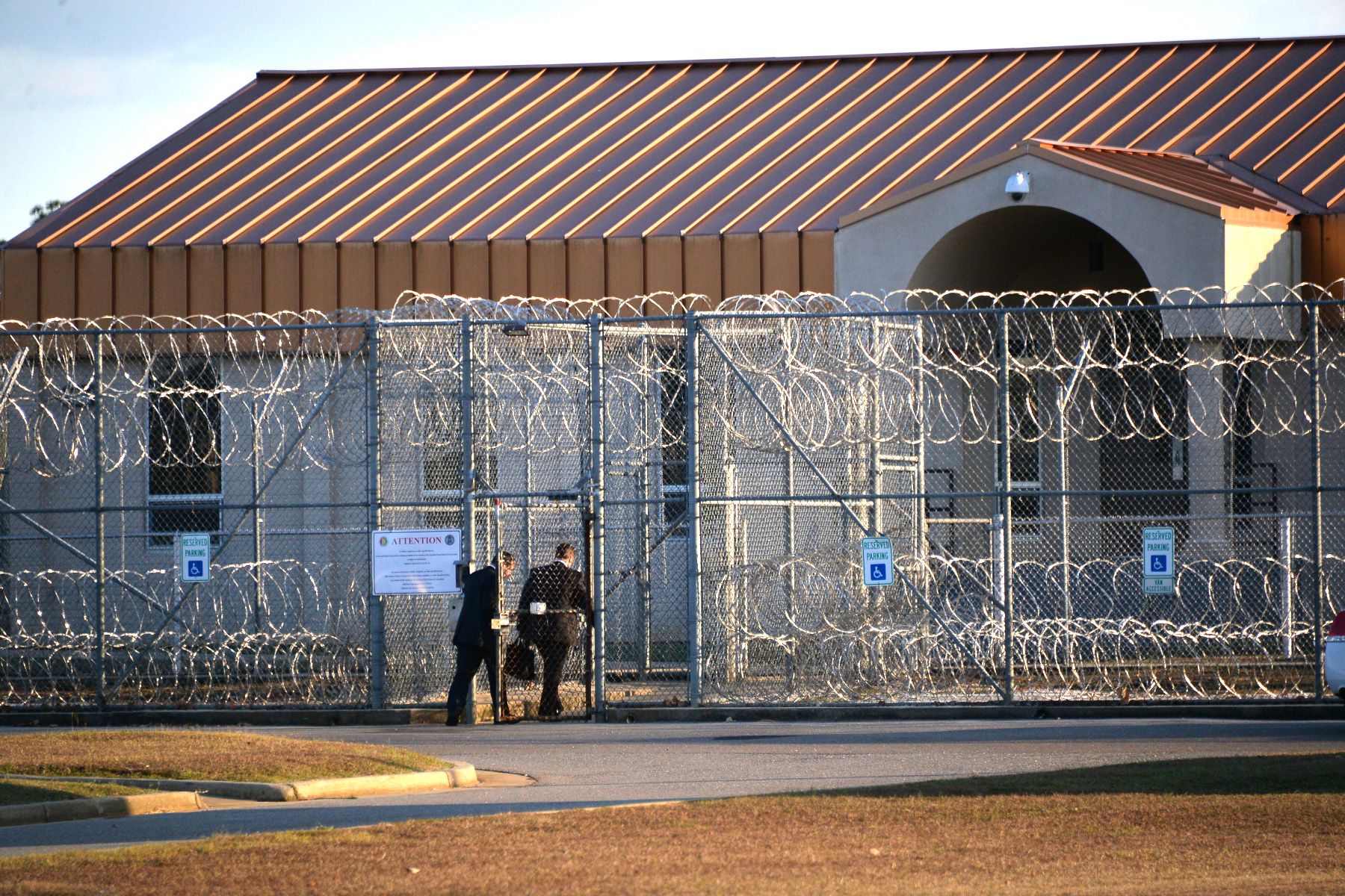 Two individuals in black suits walk through the entrance of a barbed wire fence. A tan prison is behind the fence. 