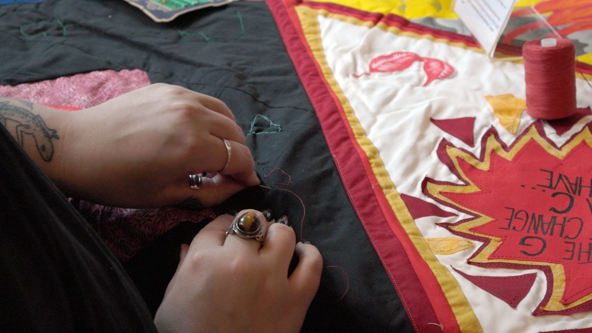 A photo shows a person's hands sewing on a piece of a quilt. On the right is a quilt patch that is red, yellow and cream, and on the left is a black piece of fabric.