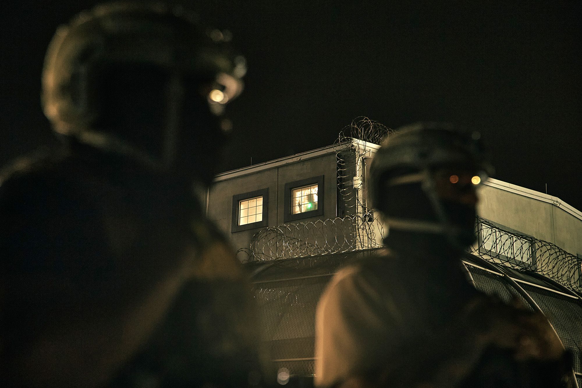 A silhouette of a person looks out of a window of a detention facility surrounded by barbed wire. Two agents with helmets and protective gear stand in the foreground. 