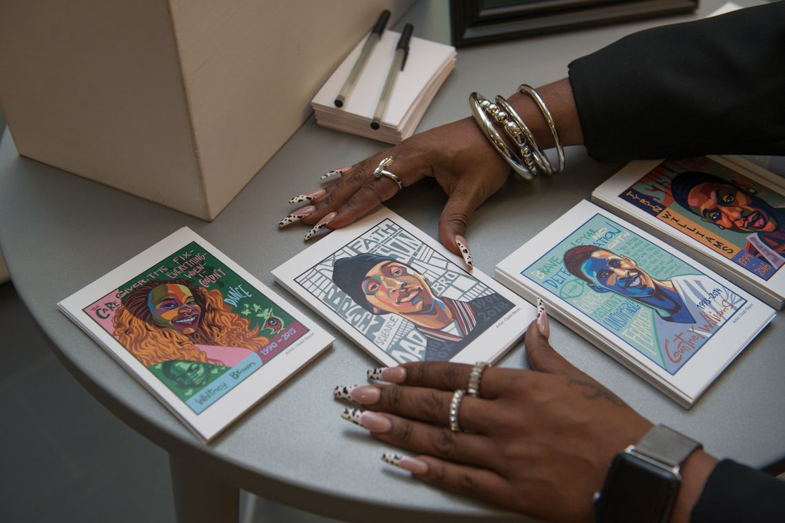 A photo of a Black woman’s hands frame a card featuring a small painted portrait of Mario Fox, a Black man wearing a hat and a bomber jacket. Next to it are three other painted portraits of, from left, Whitney Brown, Courtney Williams and Tyrin Williams. 
