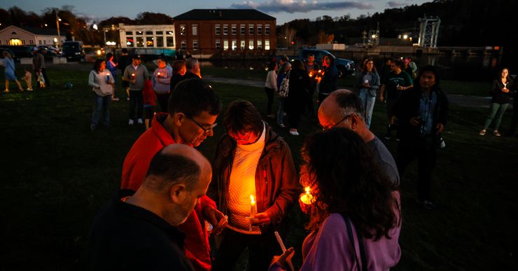 Five people in the foreground of a photo gather outdoors, holding candles.  In the background, more people hold candles in front of an event center.  