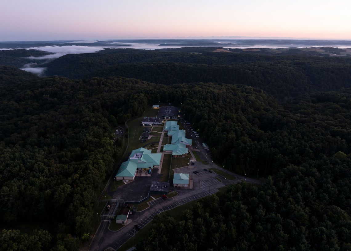 An aerial photo shows the campus after sunset. The facility has a light green roof and is surrounded by forest and hills.