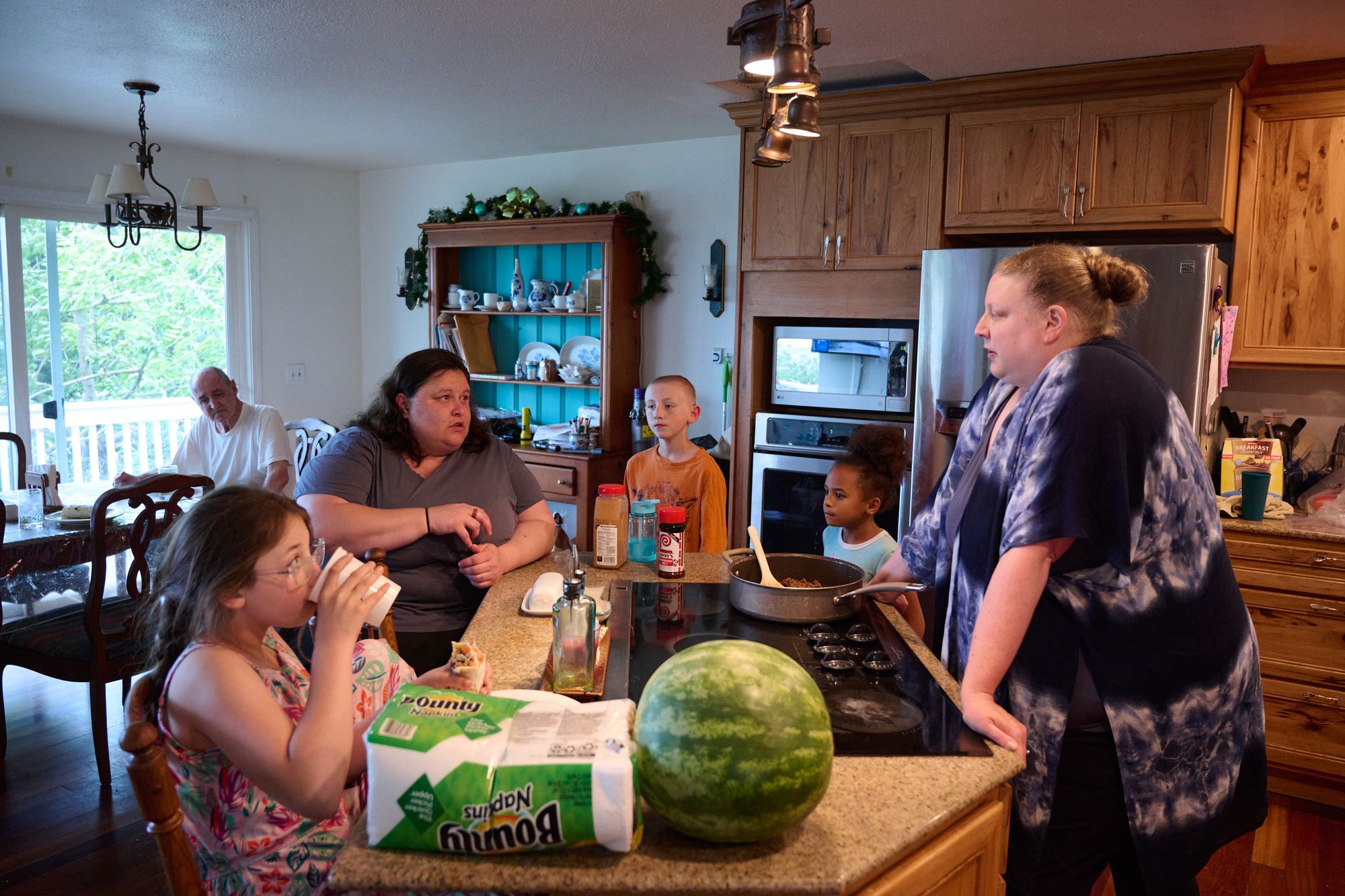 A woman with light-toned skin sits at a kitchen counter with a young girl with light-toned skin, a young boy with light-toned skin, a young girl with medium-toned skin, and another woman with light-toned skin. An older man with light-toned skin sits behind them at a table. 