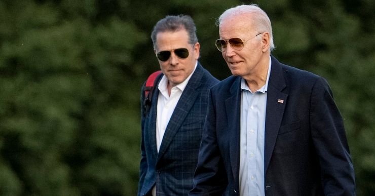 Two White men wearing sunglasses and navy suits are walking in a field. 