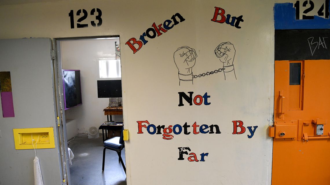 A cell door stands open next to a wall with the words "broken but not forgotten by far" painted on. A drawing of shackled hands making fists sits in the middle of the words.