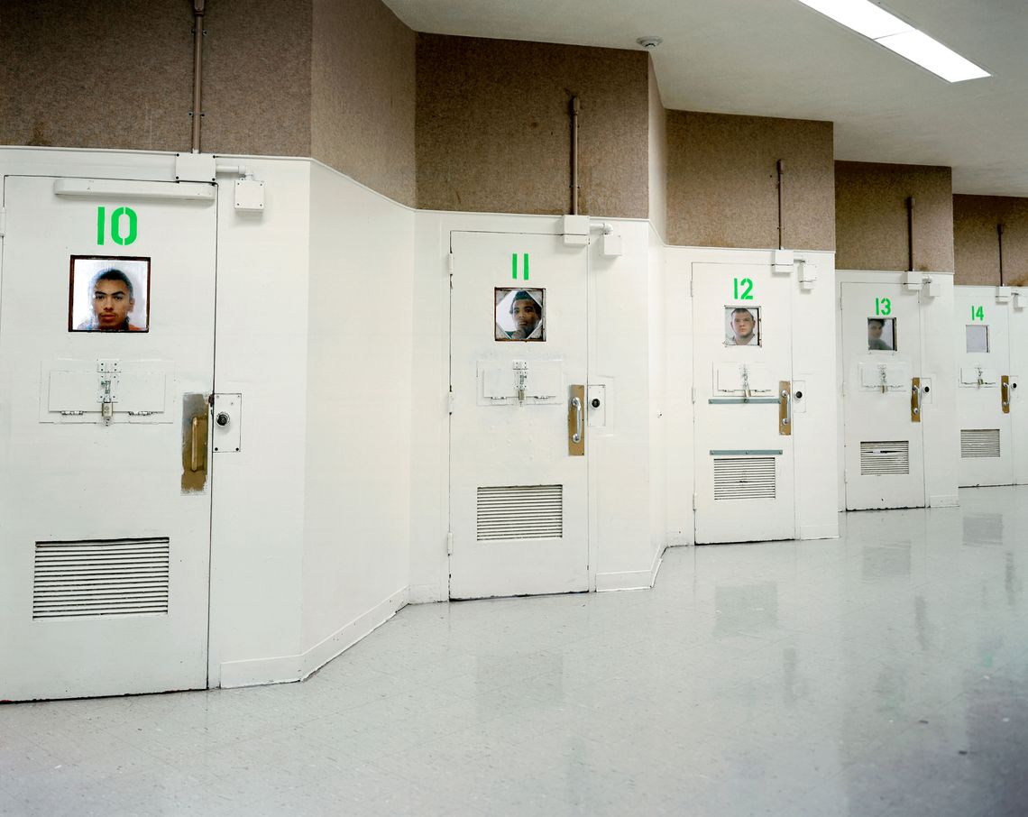 A photo shows five doors staggered down an indoor wall. In each door, there is a small square window. Through four of the windows, the face of an incarcerated young person is visible; because of the angled design, they can’t see each other. 