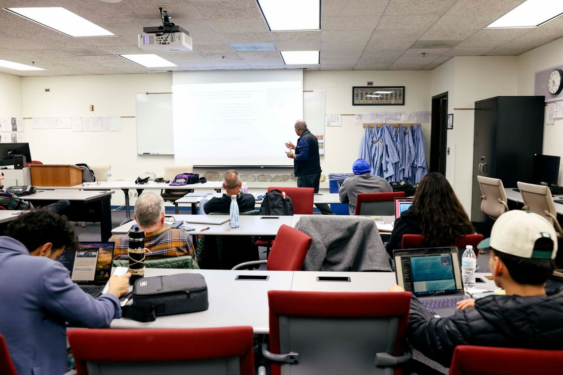 A photo shows Martinez teaching and standing in front of a class where seven students are visible in three rows. Some students have their laptops open.