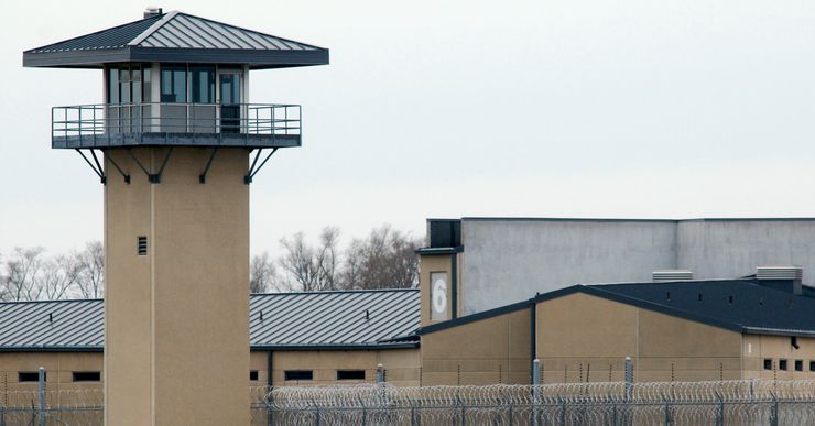 A tan guard tower and prison yard at an Illinois facility. There are wire fences and buildings in the background. 