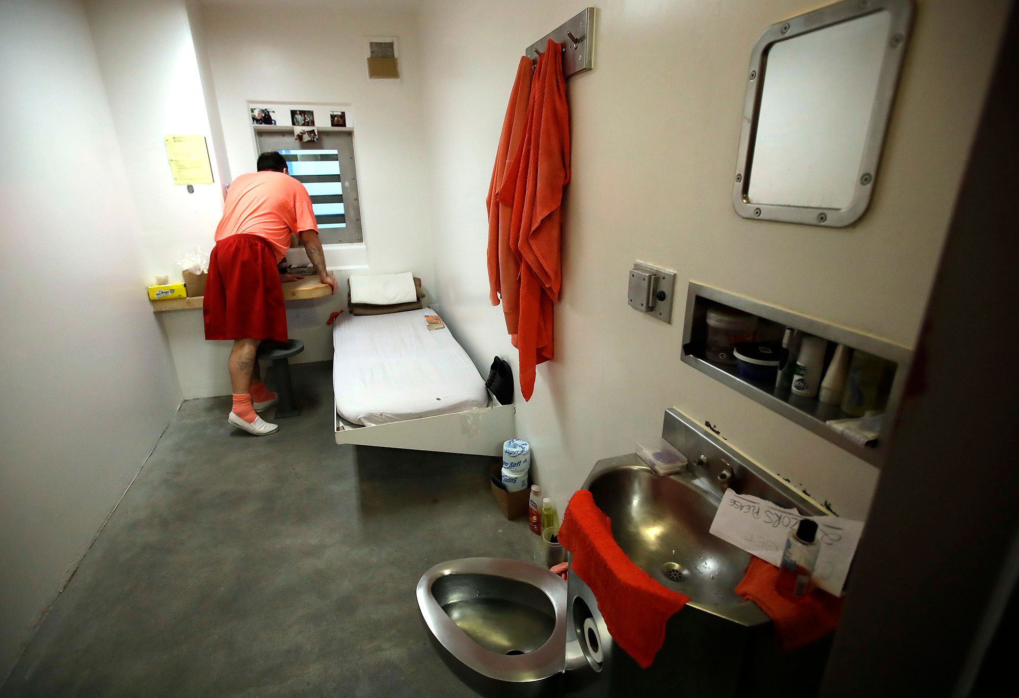 A man wearing an orange and red prison uniform looks out the window of his solitary confinement cell. 