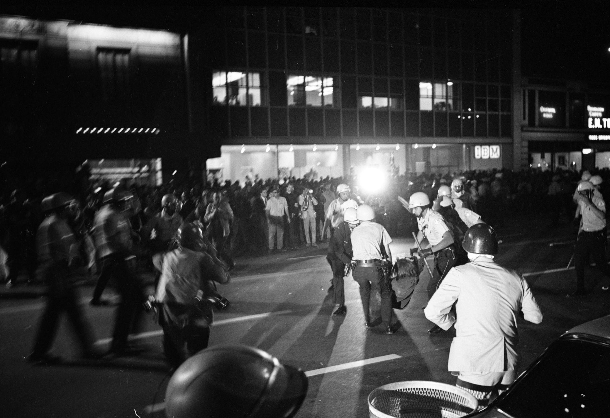 A black and white photo shows a group of officers carrying a protester. In the background, a large crowd of protesters is on the street. 