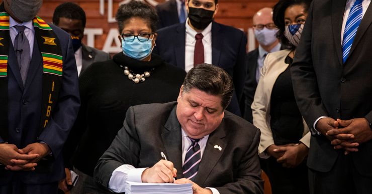 Gov. J.B. Pritzker, a seated man with light-toned skin and short-cropped dark hair, signs a bill that is composed of a high stack of papers. Eight masked people with varying skin tones and genders stand behind him.