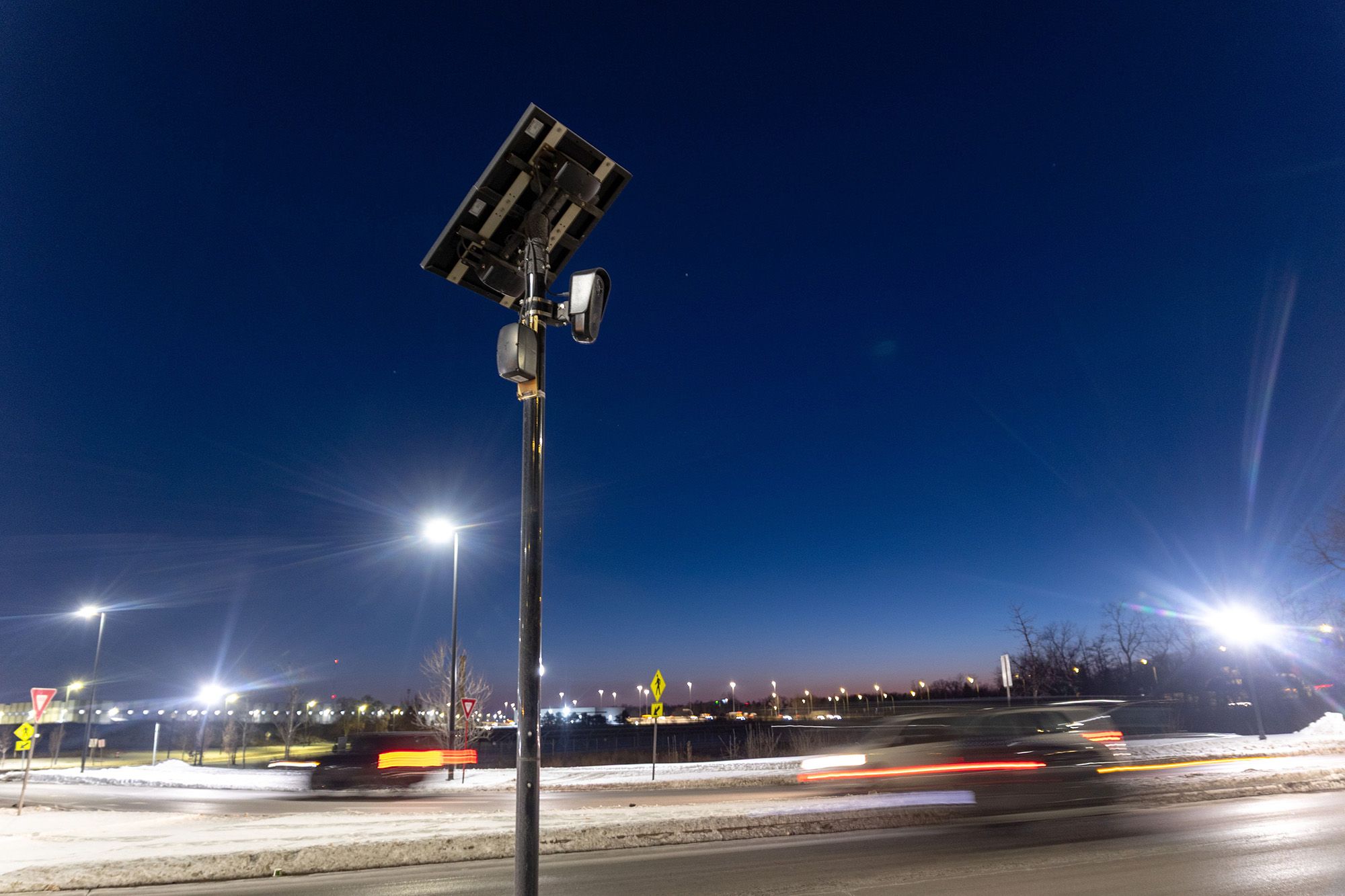 A photo shows cars driving at night in a blur past a Flock camera, which is situated at the top of a pole. There is a square plate at the top of the pole. 