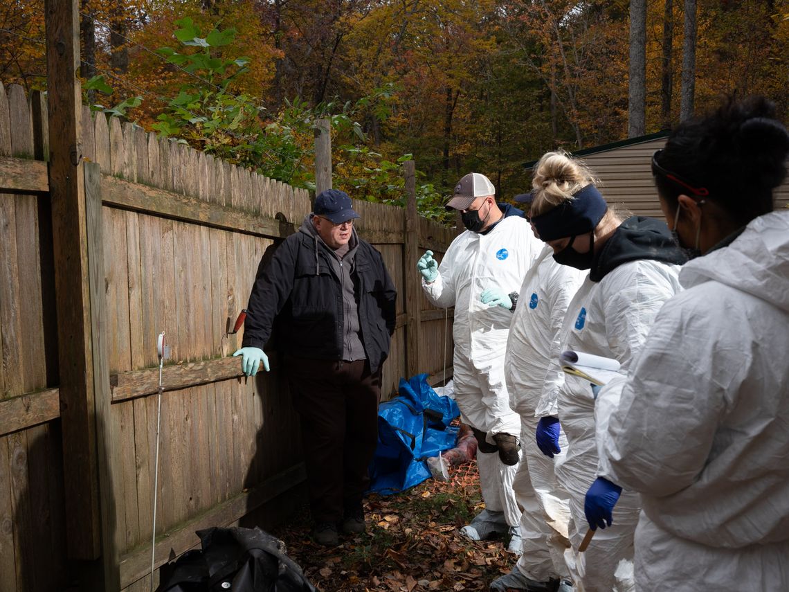 Students wearing white hazmat suits stand to the right of Pat Cicero, an instructor, who is clothed in a black jacket and gray hooded sweatshirt. 