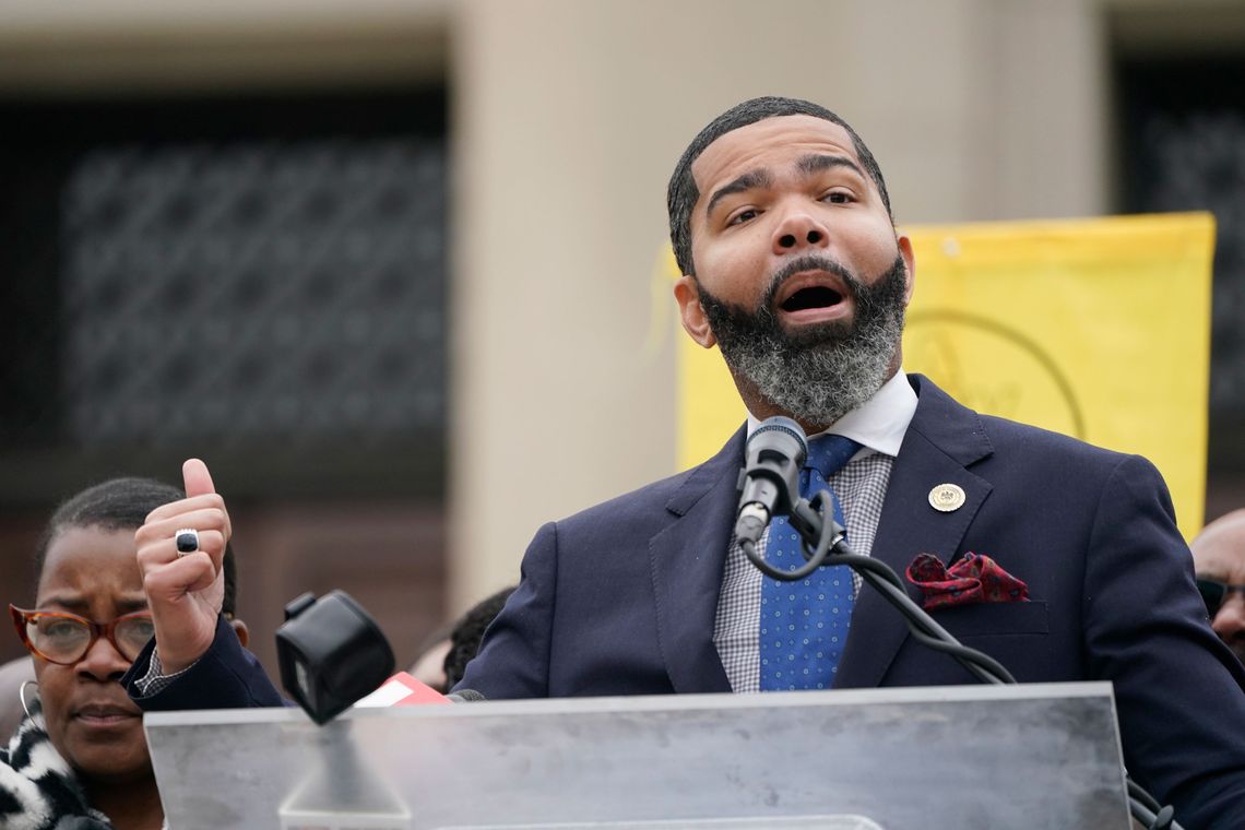 A middle-aged Black man wearing a suit with a blue tie speaks at a podium.